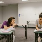 Two college students in a classroom setting, one appears to be copying from the other's paper.