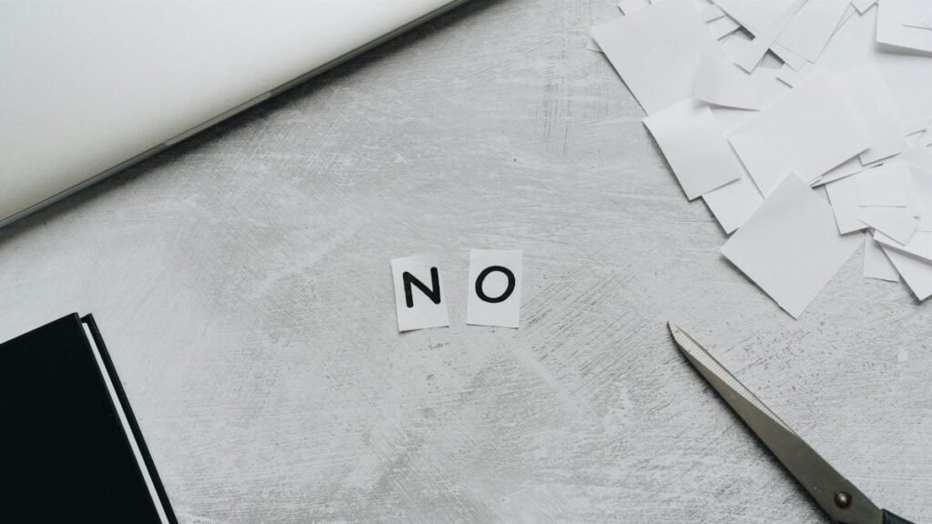 Top view of scattered paper squares, laptop, and scissors forming the word 'NO', implying rejection or denial.
