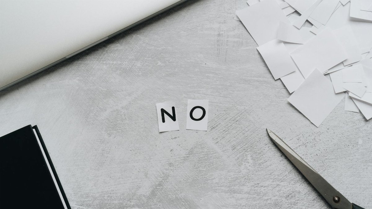 Top view of scattered paper squares, laptop, and scissors forming the word 'NO', implying rejection or denial.