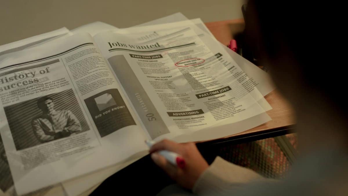 Person reviewing job listings in a newspaper, holding a pink marker for highlighting job ads.