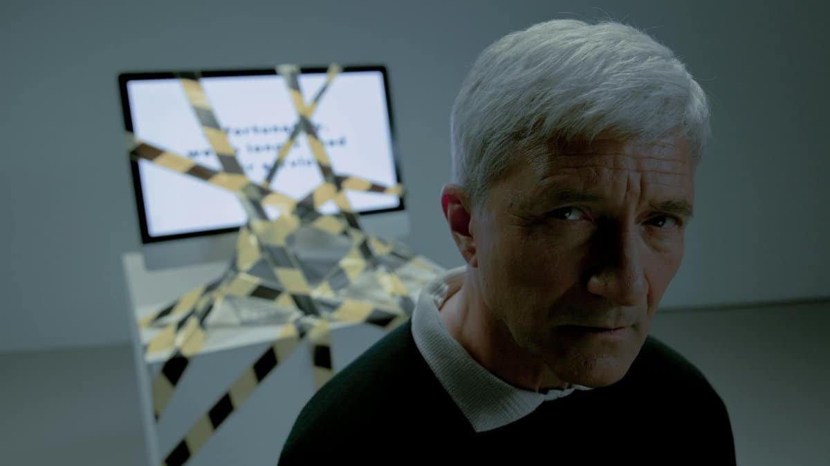 A senior man looks concerned, standing near a computer covered in caution tape, symbolizing job uncertainty.