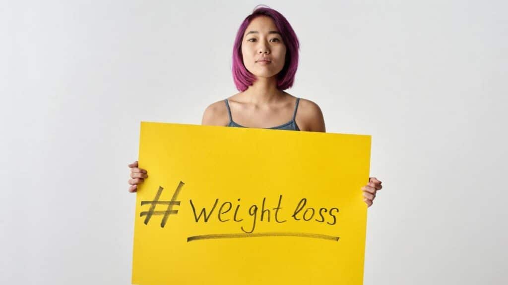 Asian woman with purple hair holding a weight loss sign in a studio setting.