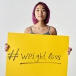 Asian woman with purple hair holding a weight loss sign in a studio setting.