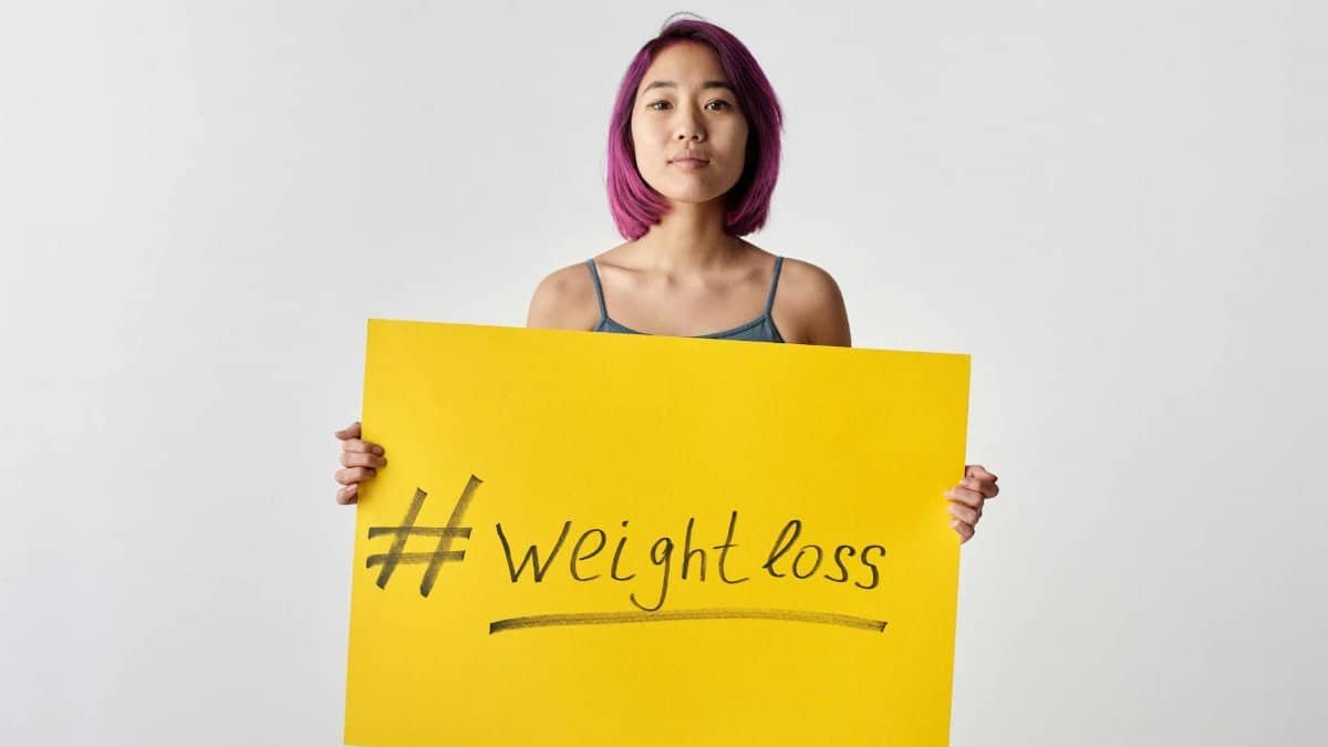 Asian woman with purple hair holding a weight loss sign in a studio setting.