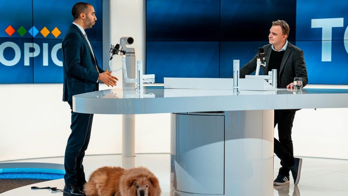 Two hosts discussing at a modern TV studio with a dog laying by. Engaging professional setup.