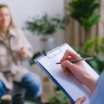 Unrecognizable professional female psychologist writing on clipboard while sitting against client on blurred background during psychotherapy session in light office