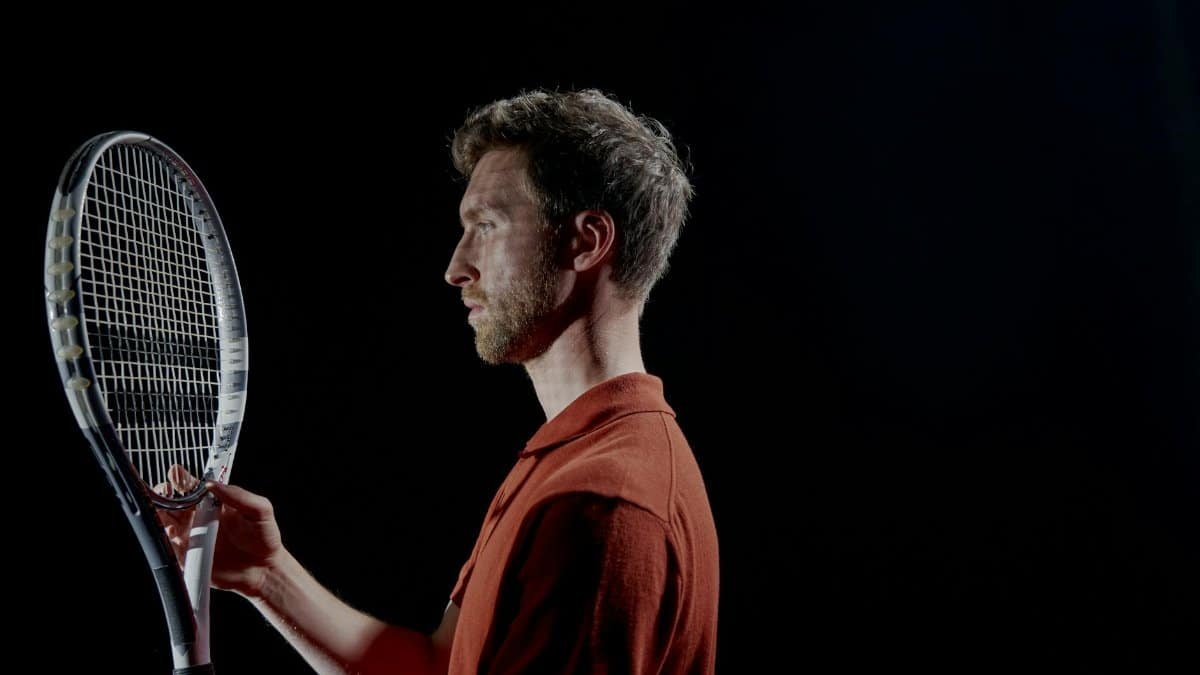Profile view of a focused male tennis player holding a racket in a dimly lit setting.