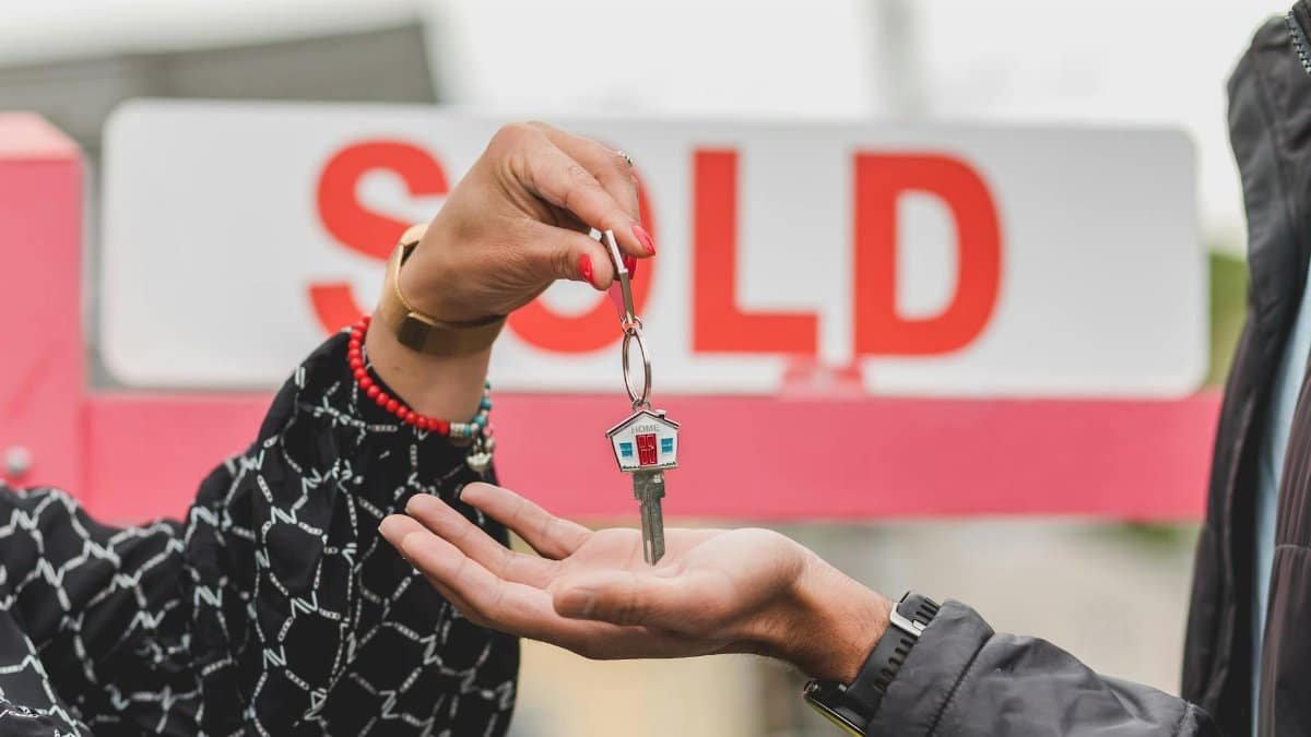 Close-up of handing over house key with sold sign in background, symbolizing real estate success.