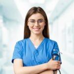 Young nurse in blue scrubs smiling and holding a stethoscope in a hospital corridor.
