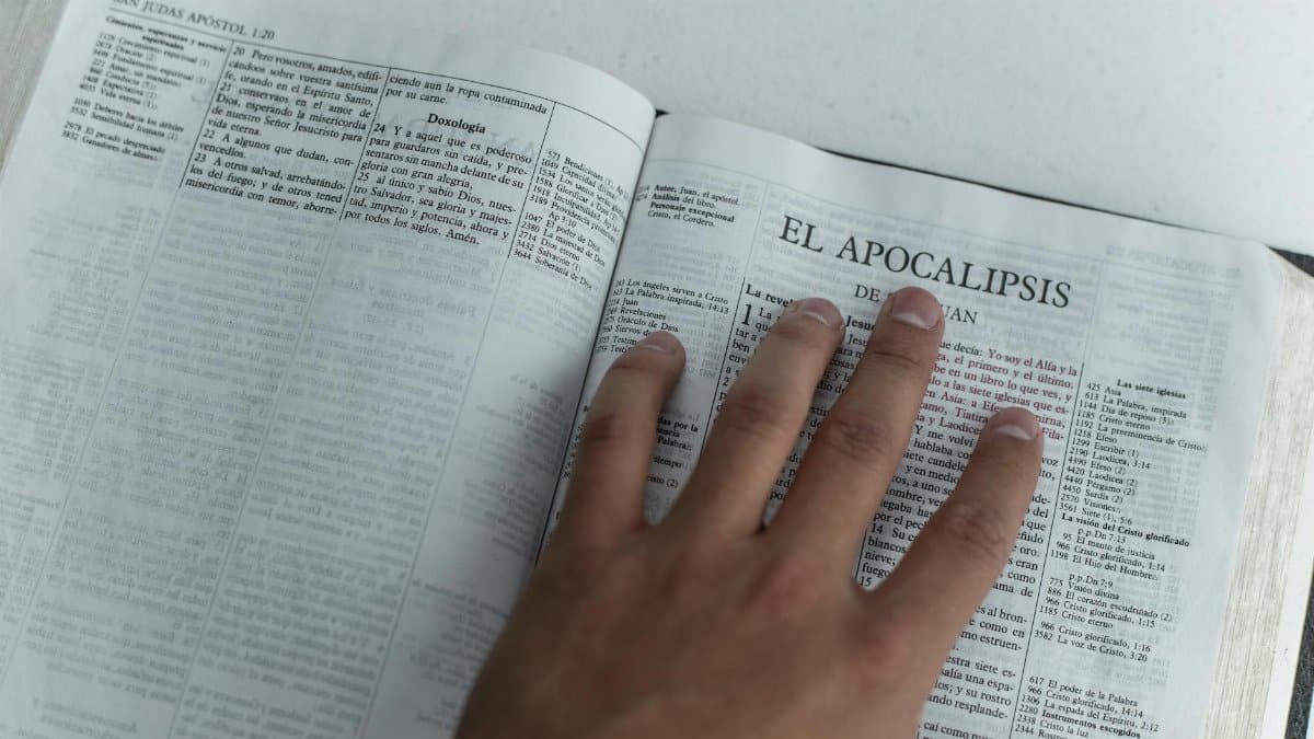A hand touching Bible pages open to the Book of Revelation, symbolizing faith and study.
