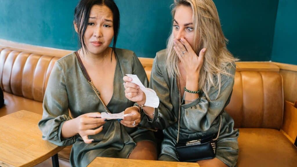 Two women express surprise while reviewing a restaurant bill in a casual setting.