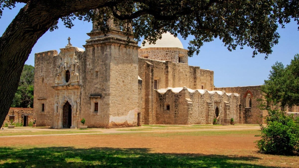 Statue of an old mission with lush trees in San Antonio, capturing historical architecture.