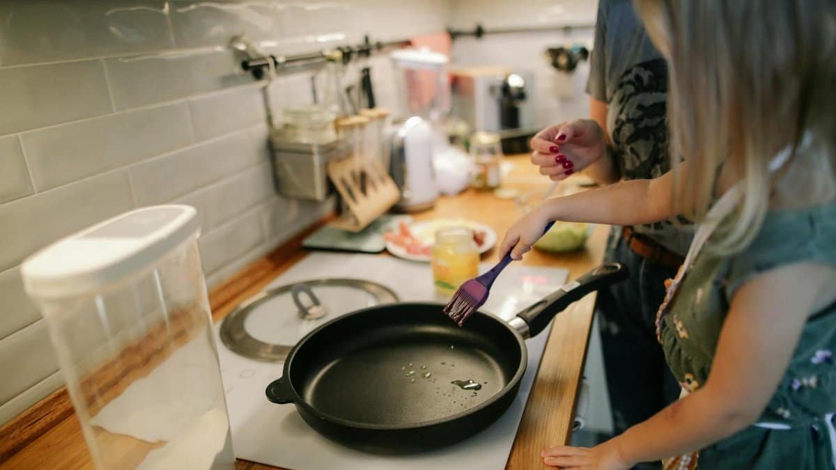 Young girl cooking with a frying pan under adult supervision in a modern kitchen.
