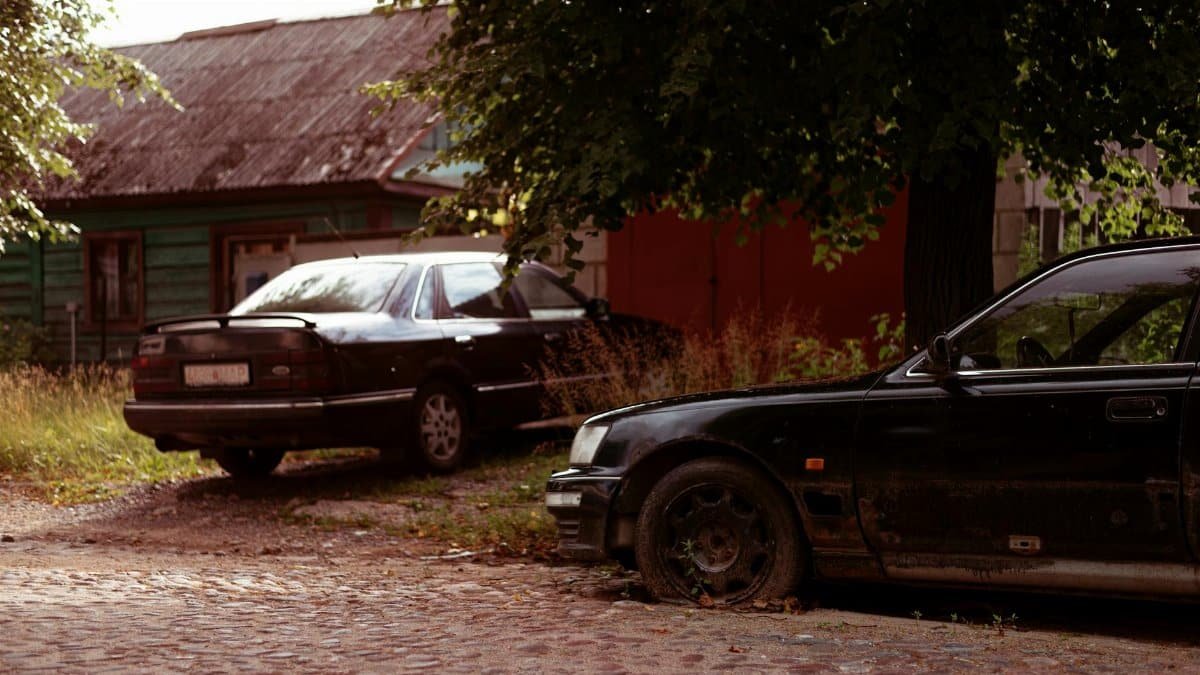 Two black cars in a neglected suburban driveway beside an old house.