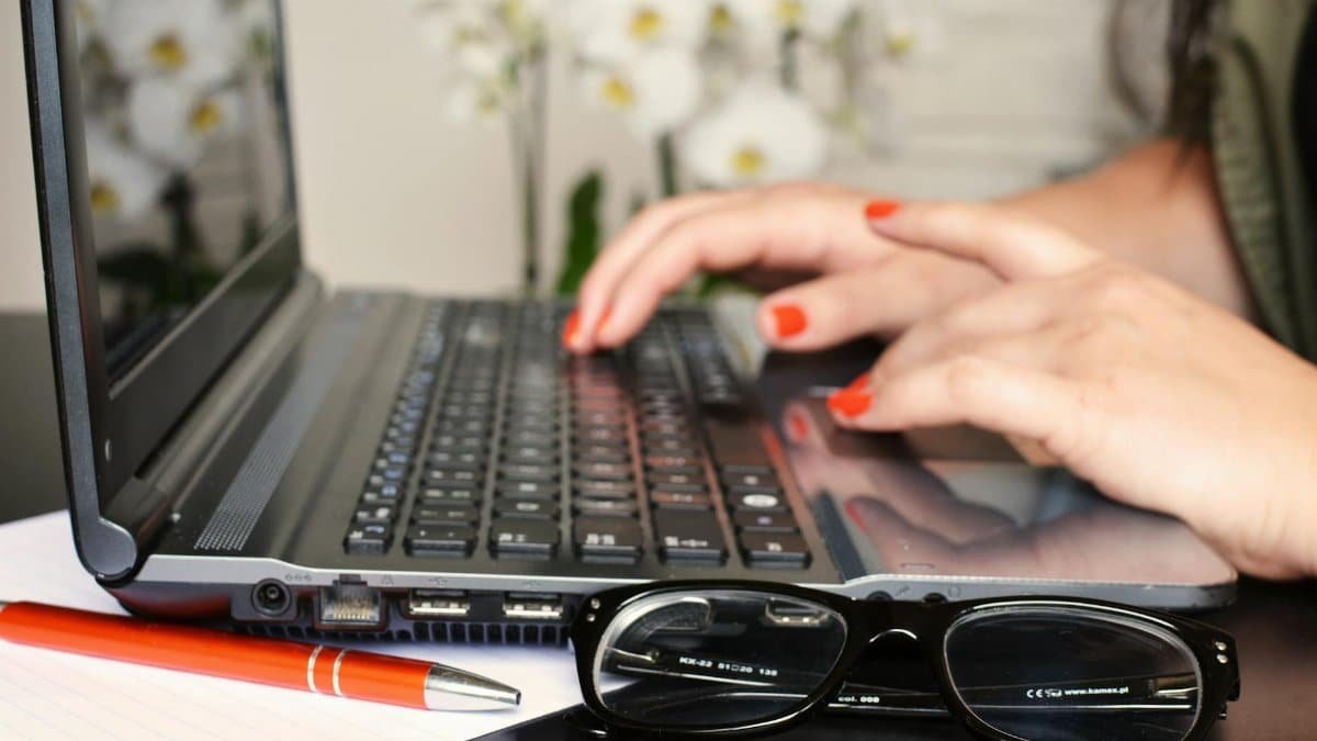 Close-up of a woman's hands typing on a laptop with glasses and a notepad on the desk.