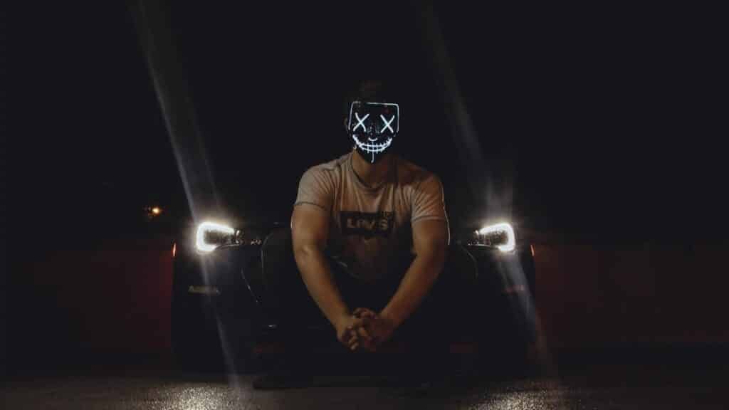 A man wearing an illuminated LED mask sits in front of a car with headlights on at night.