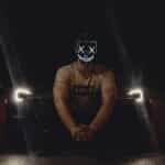 A man wearing an illuminated LED mask sits in front of a car with headlights on at night.