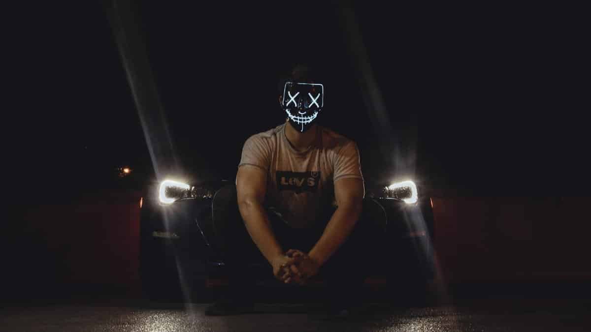 A man wearing an illuminated LED mask sits in front of a car with headlights on at night.