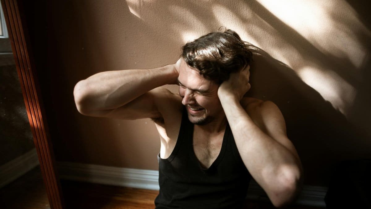 A man sitting on the floor indoors, showing signs of stress by covering his ears.