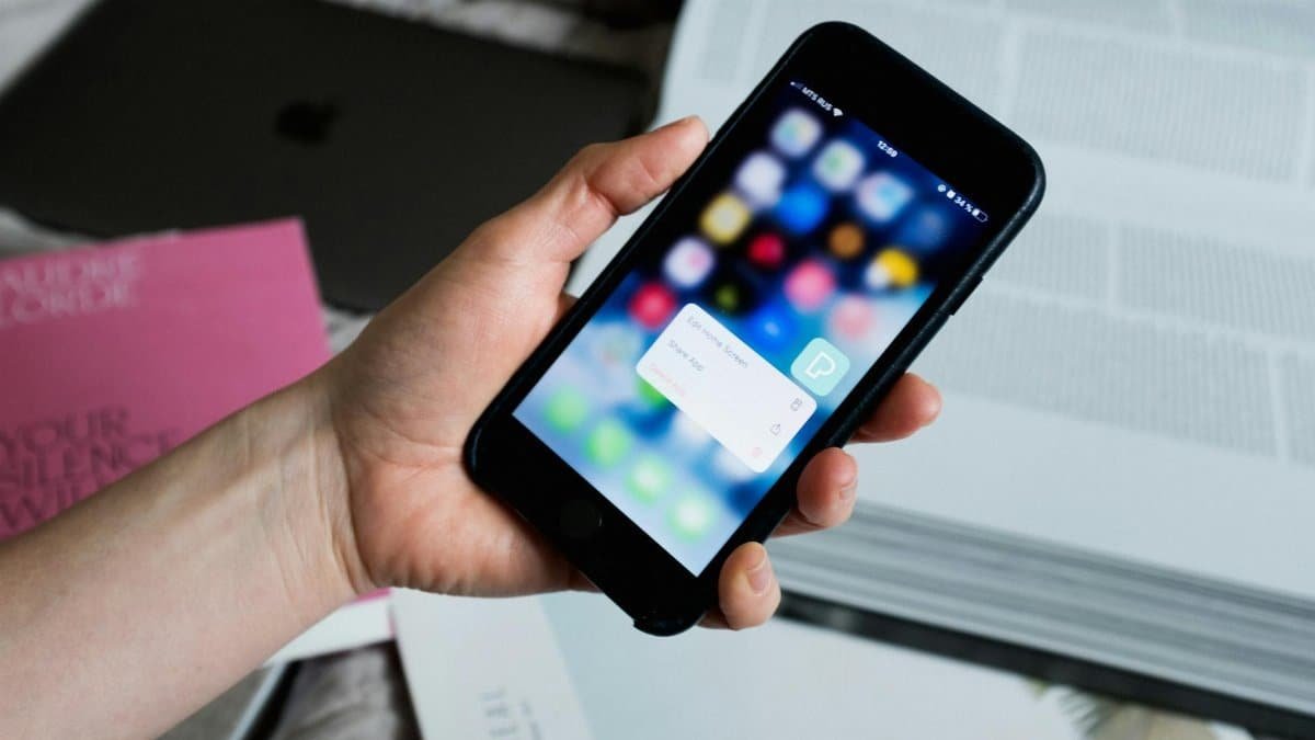 From above of crop anonymous person taking cellphone while demonstrating pop up window on blurred screen with application logos and having books on bed sheet