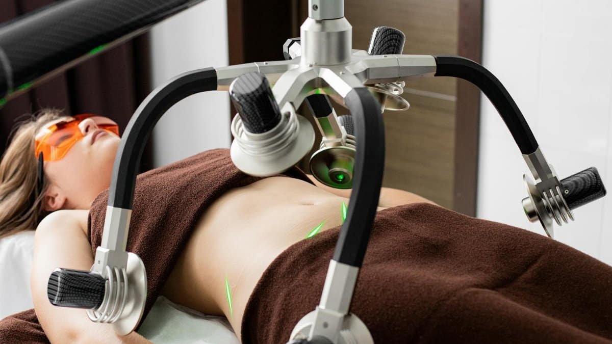 Female client lying on table under lasers of modern weight loss machine on belly during liposuction procedure in medical clinic