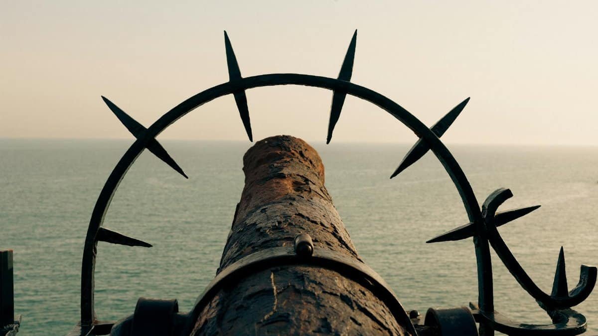 A rusted cannon aimed towards the sea, framed by metal spikes, suggesting a historic maritime setting.