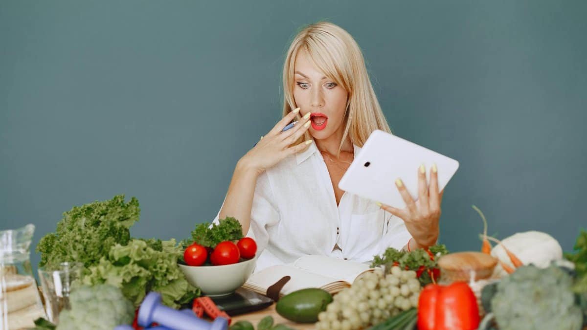 A surprised woman looking at a tablet surrounded by fresh vegetables and fruits, promoting healthy eating.