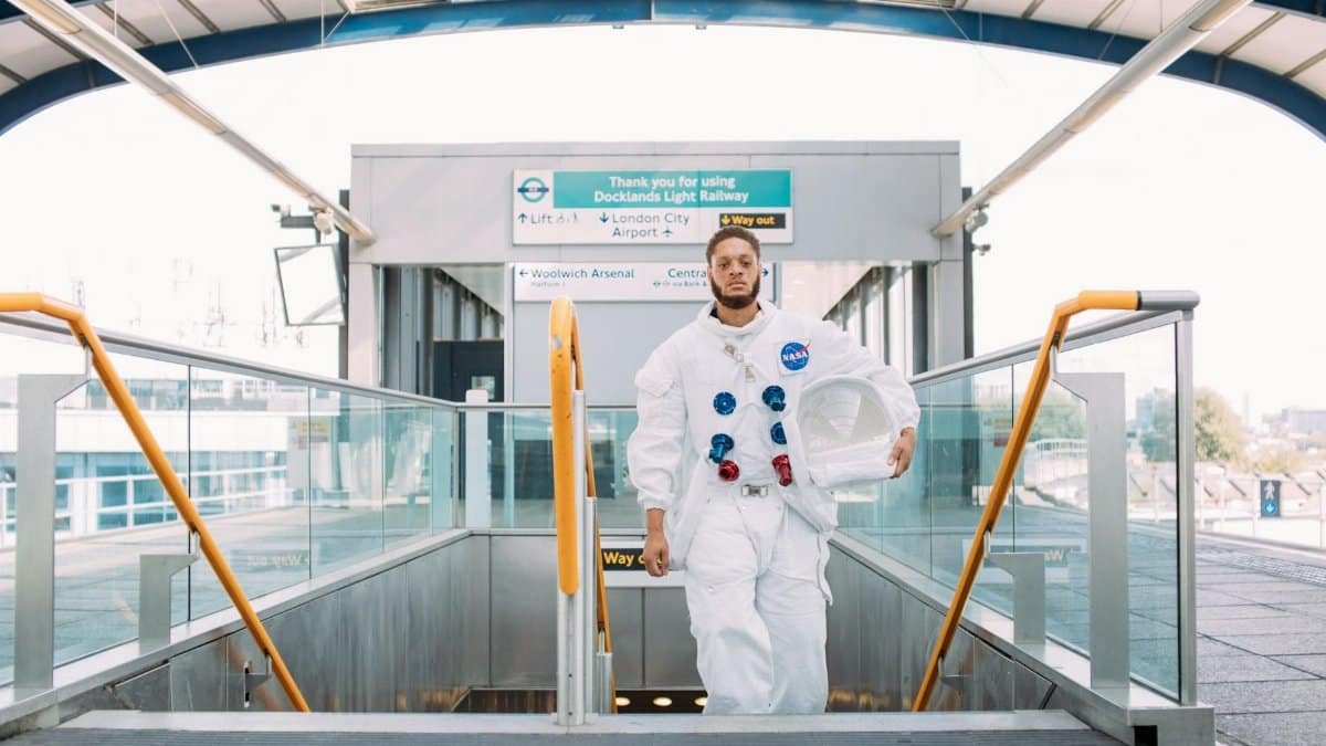 A man dressed as an astronaut at a London DLR station, holding a helmet.