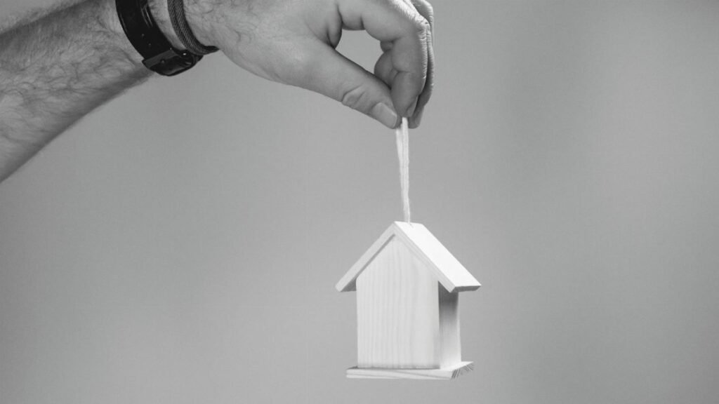 Black and white photo of a hand holding a miniature wooden house by a string, symbolizing real estate.