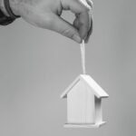 Black and white photo of a hand holding a miniature wooden house by a string, symbolizing real estate.