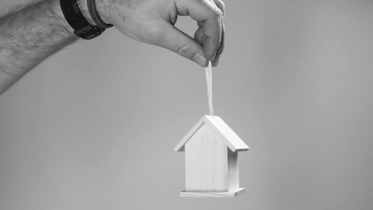 Black and white photo of a hand holding a miniature wooden house by a string, symbolizing real estate.