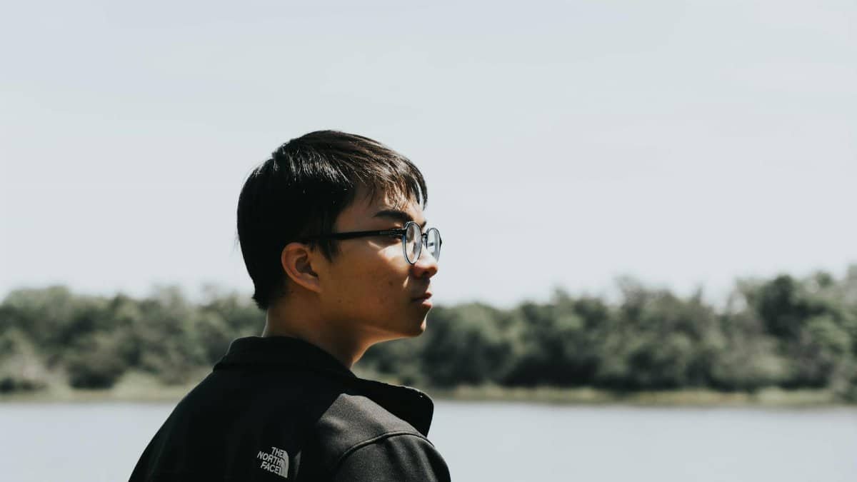 Teenager wearing glasses looking across a serene lake with trees in the background.