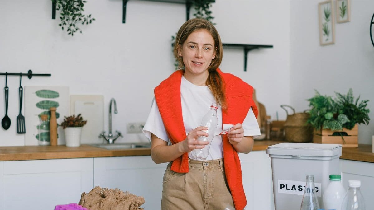 Female holding plastic bottle and looking at camera while sorting trash in blurred kitchen