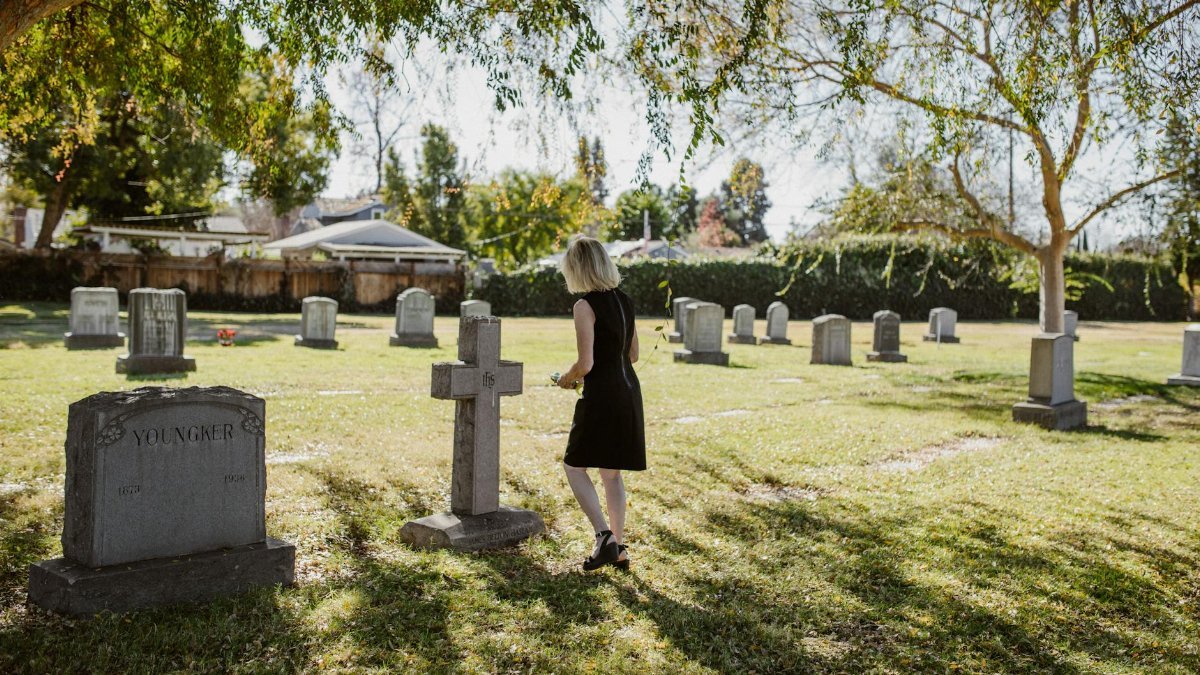 Woman in black dress walking among gravestones in a sunny cemetery setting.