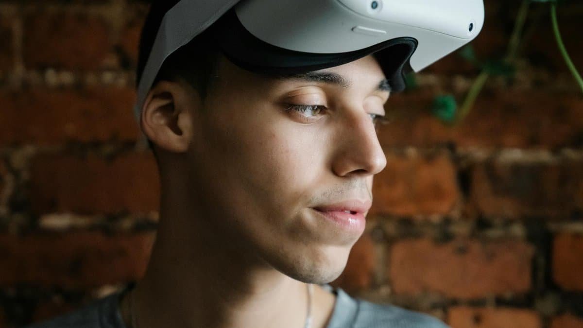 Portrait of a young man wearing a virtual reality headset, looking thoughtful against a brick wall indoors.