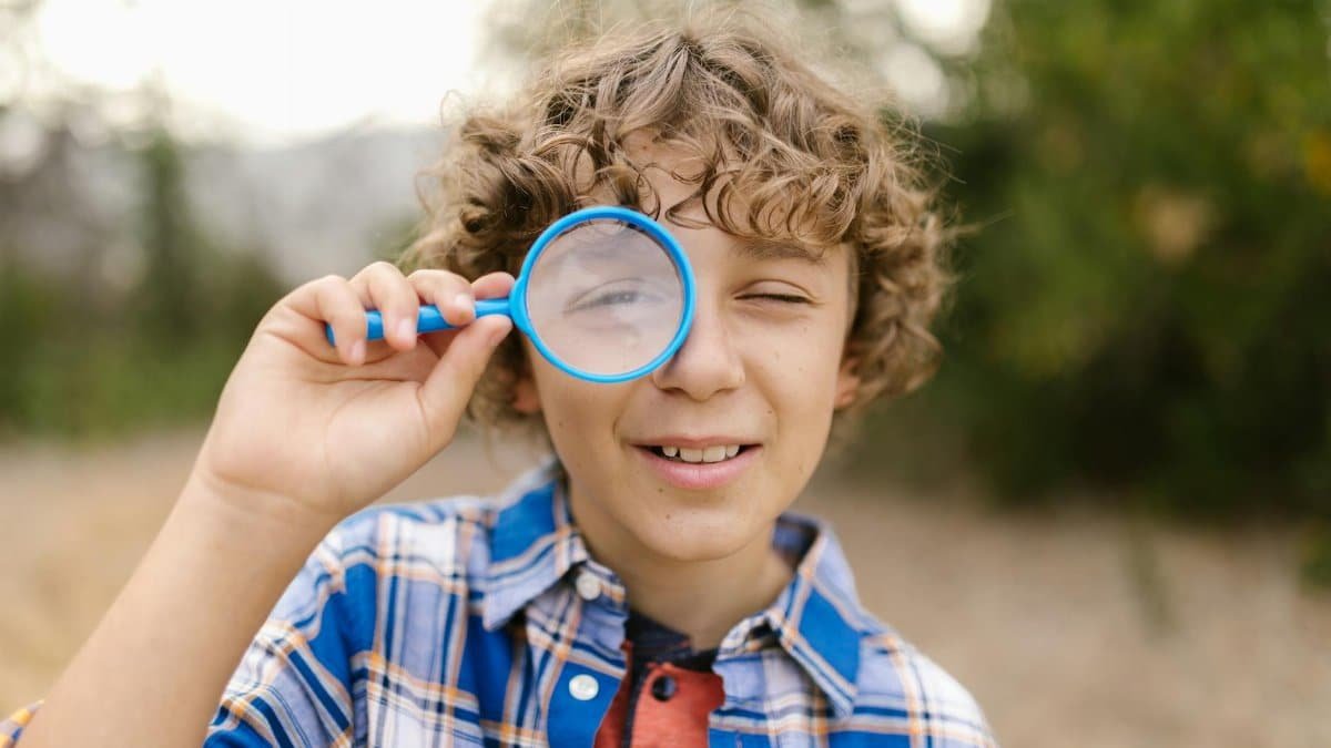 A happy child with curly hair looking through a magnifying glass in a playful outdoor setting.