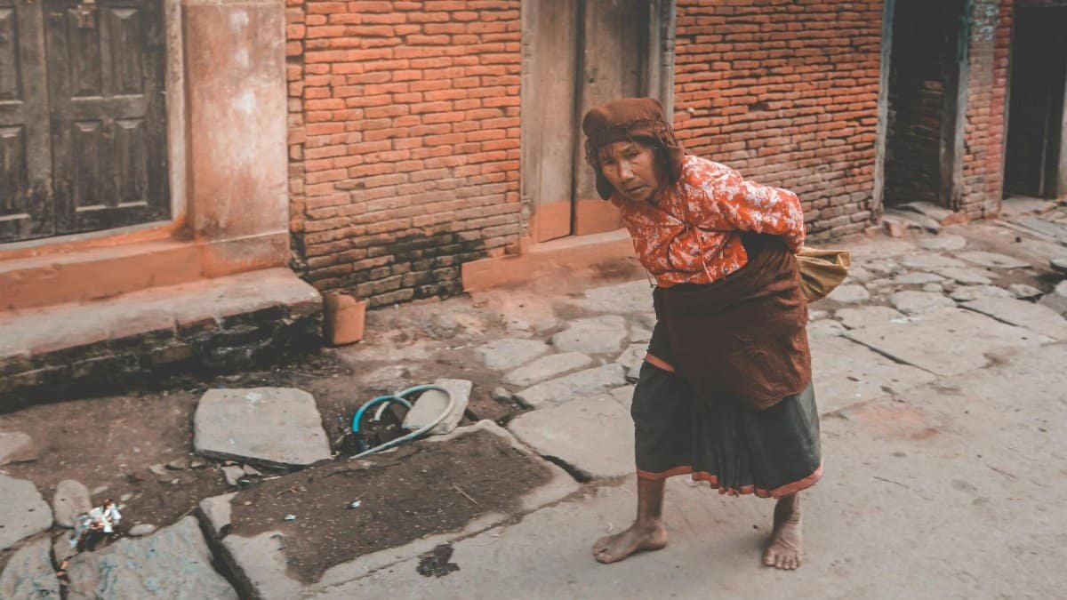 From above of mature barefoot ethnic female leaning forward while looking away near old brick house in town