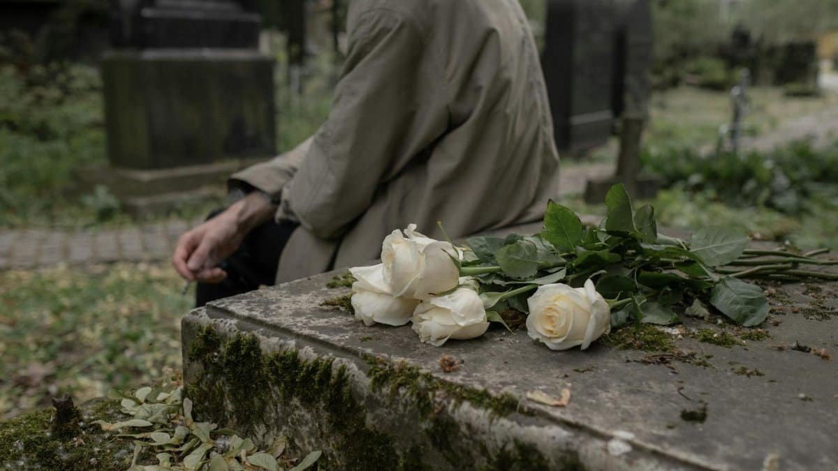 A person sitting by a tombstone with white roses, evoking themes of grief and remembrance.