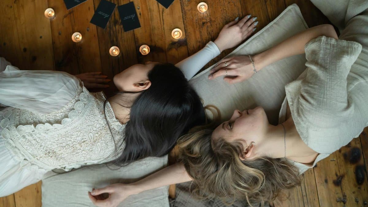 Two women lying on cushions with candles and tarot cards on wooden floor, creating a peaceful atmosphere.