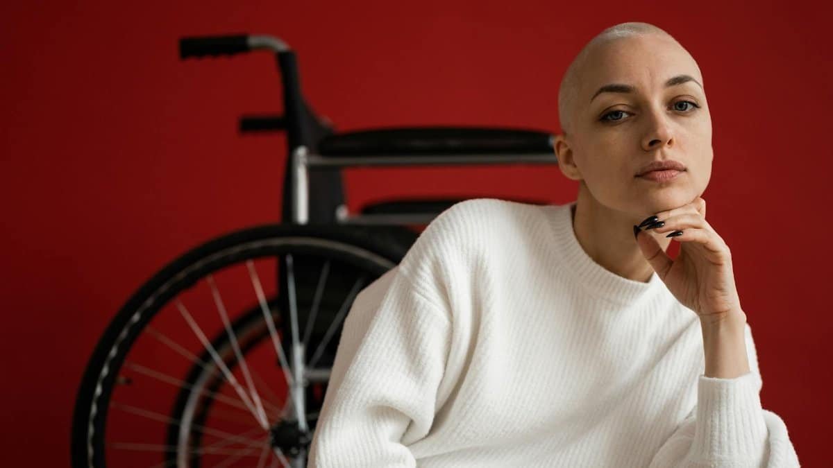 Portrait of a confident woman in white sweater sitting near a wheelchair against red background.