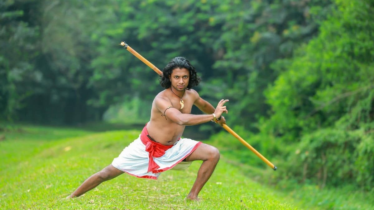 Asian man practicing Silambam martial arts outdoors with a bamboo stick.