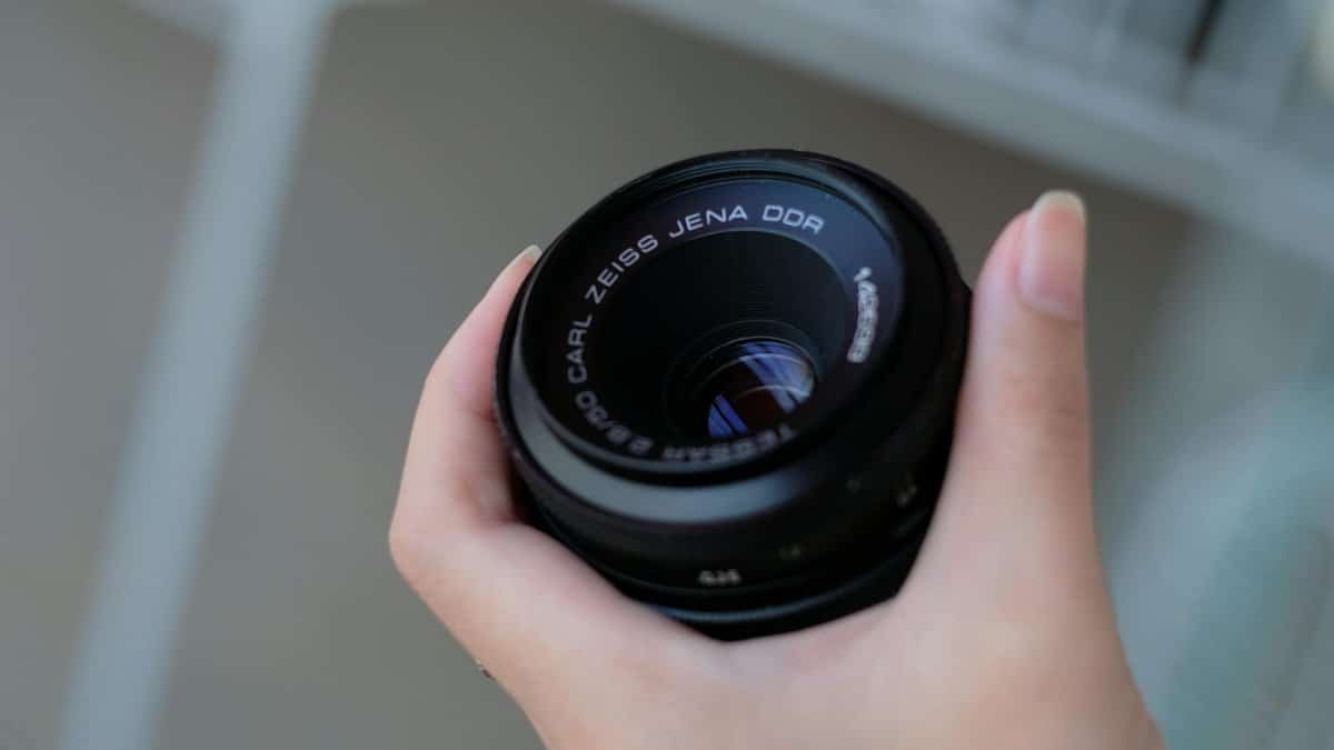 Close-up of a hand holding a Carl Zeiss Jena DDR camera lens with a blurred background.