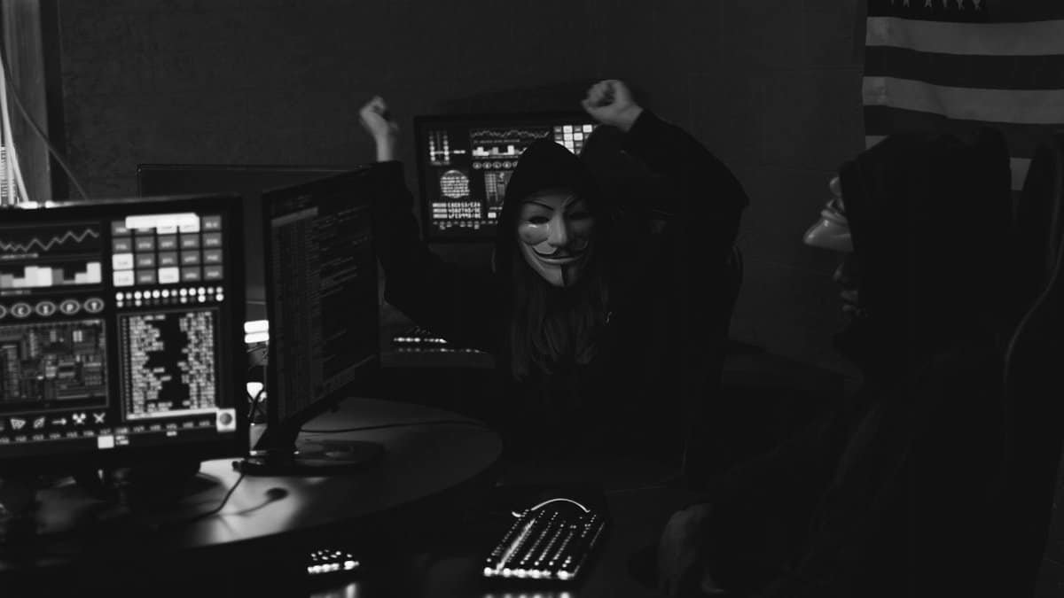 Hacker group with masks celebrating a successful cyber attack in a dimly lit room.