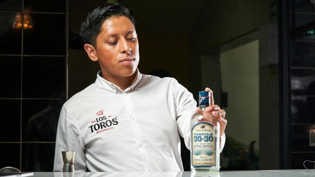 A bartender presents a bottle of Tequila 30-30 in a restaurant environment.