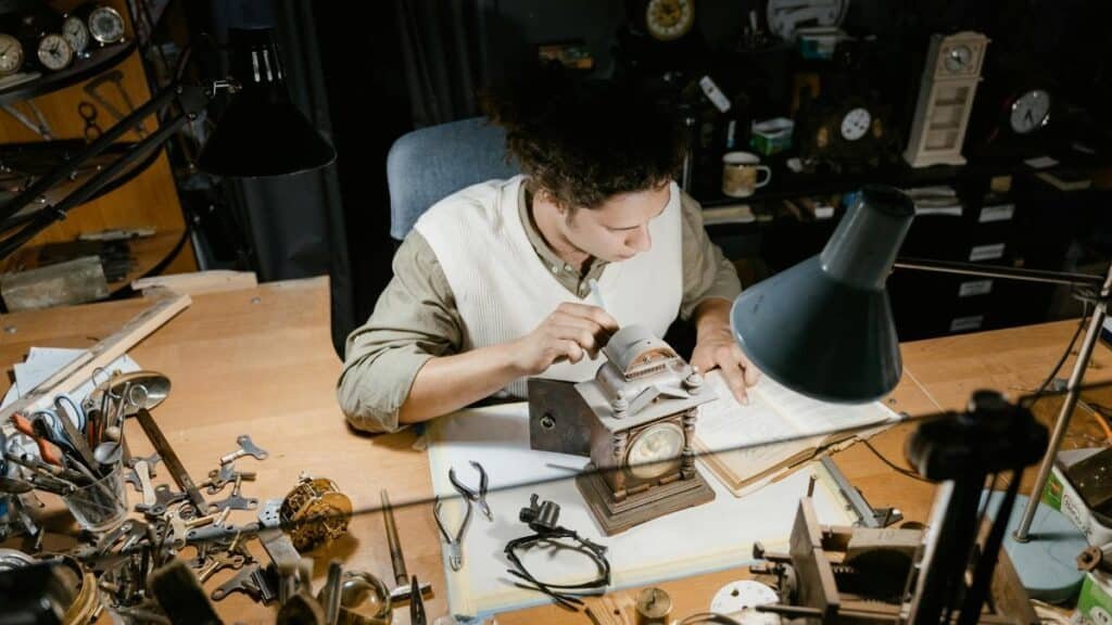 A watchmaker carefully repairs a clock on a cluttered workbench surrounded by tools.