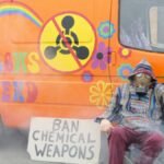 A protester in a gas mask sits by a colorfully painted van with an anti-chemical weapons sign.