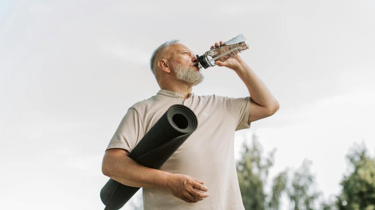 Elderly man drinking water while holding a yoga mat outdoors, promoting fitness and hydration.