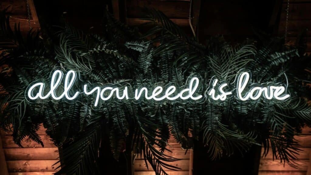 A vibrant neon sign stating 'All You Need is Love' surrounded by lush green leaves.