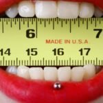 Close-up of red-lipsticked mouth holding a yellow tape measure horizontally, demonstrating measurement concept.