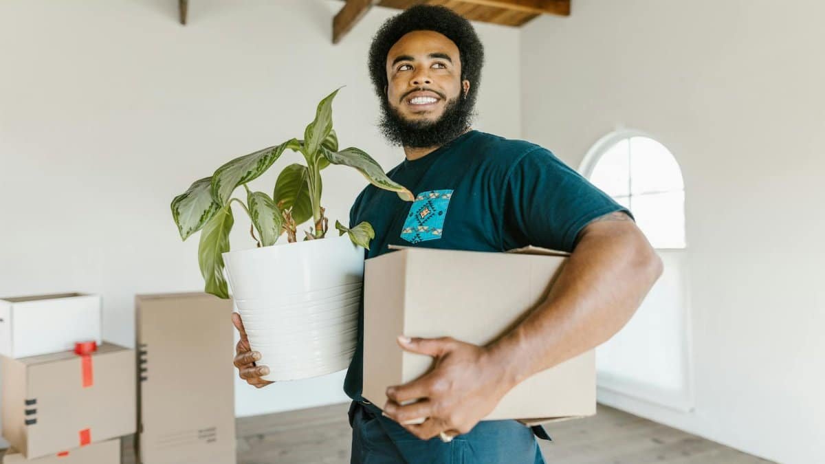 Smiling man holding a moving box and potted plant in a bright new home.